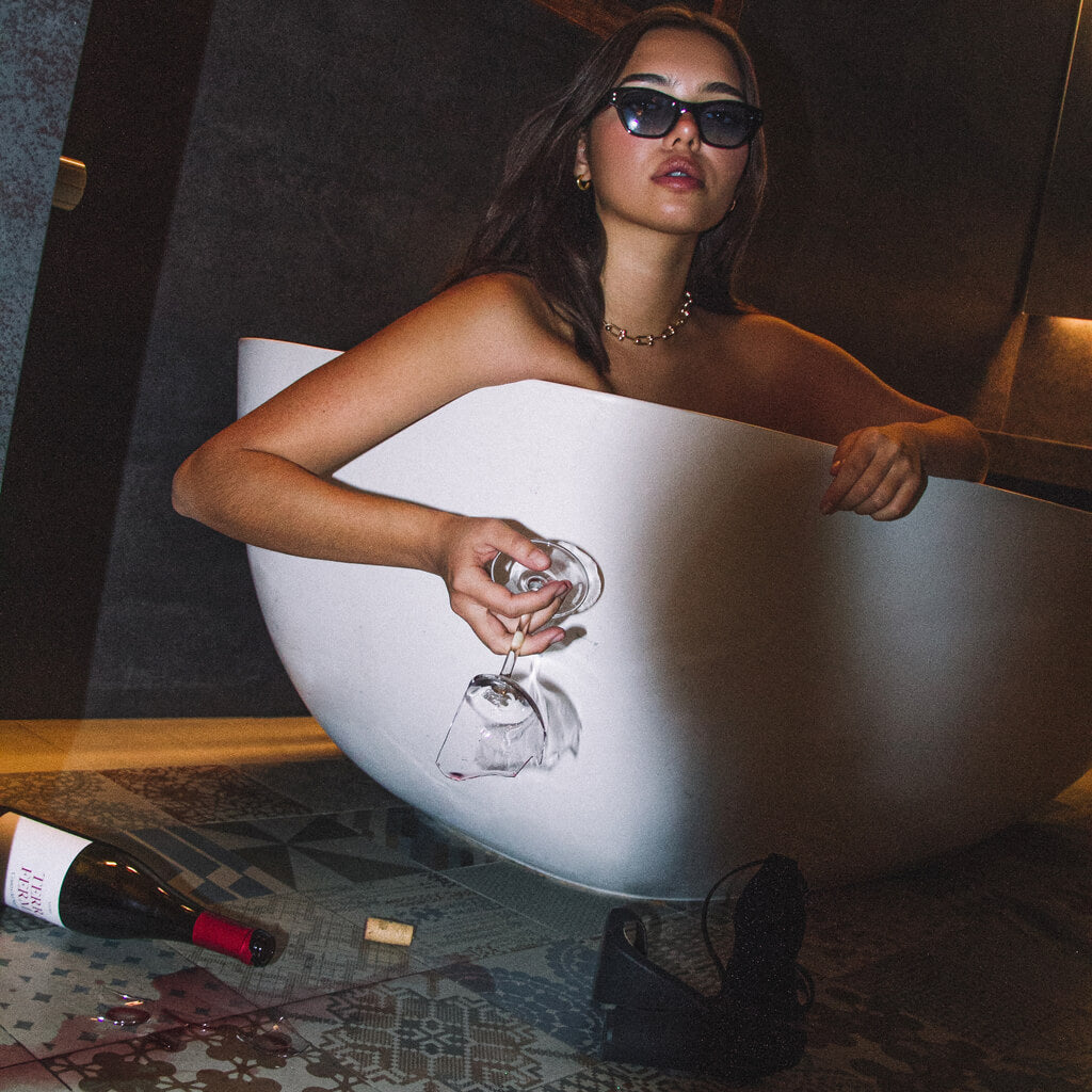girl laying in bath tub holding a broken glass of wine in vega